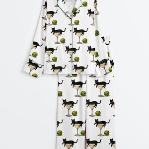 May include: White pyjama set featuring a repeating pattern of black cats in martini glasses and green olives. The long-sleeved top and trousers have green trim. The pyjamas are made of a soft, comfortable material.