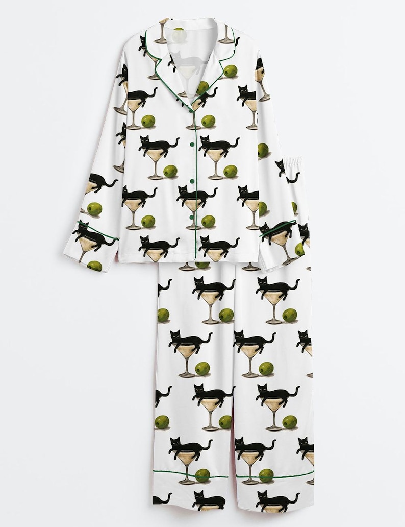 May include: White pyjama set featuring a repeating pattern of black cats in martini glasses and green olives. The long-sleeved top and trousers have green trim. The pyjamas are made of a soft, comfortable material.
