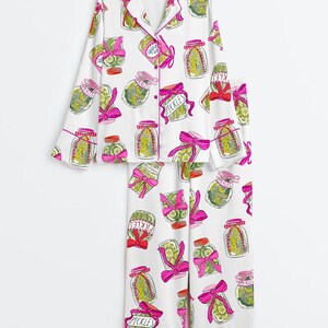 Puede incluir: Conjunto de pijama blanco con un estampado repetido de tarros de pepinillos con lazos rosas y la palabra "Pickles". La parte superior de manga larga y los pantalones tienen ribetes rosas. Los tarros contienen pepinillos verdes.