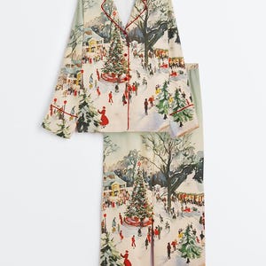 May include: A two-piece pajama set featuring a winter scene print. The set includes a long-sleeved top with red trim and a button-down front, and matching pants. The print depicts a snowy town square with a Christmas tree and people.