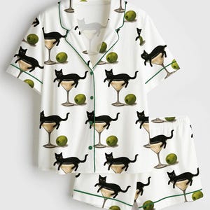 May include: White pyjama set featuring a pattern of black cats in martini glasses and green olives. The short-sleeved top and shorts have green trim.