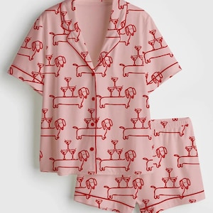 K&ouml;nnte beinhalten: Rosa Pyjama-Set mit rotem Dackel- und Cocktailglas-Muster. Das kurz&auml;rmelige Button-up-Shirt und die passenden Shorts zeigen ein sich wiederholendes Design von Hunden, die Martinigl&auml;ser balancieren. Das Set hat rote Paspeln.