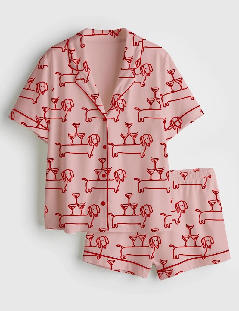 K&ouml;nnte beinhalten: Rosa Pyjama-Set mit rotem Dackel- und Cocktailglas-Muster. Das kurz&auml;rmelige Button-up-Shirt und die passenden Shorts zeigen ein sich wiederholendes Design von Hunden, die Martinigl&auml;ser balancieren. Das Set hat rote Paspeln.