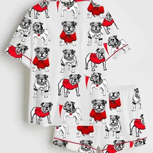 May include: White pyjama set with a repeating pattern of bulldog illustrations. The bulldogs are in black and white, some wearing red sweaters. The set includes a short-sleeved top and matching shorts, both with red trim.