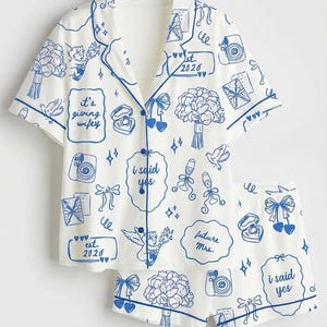 May include: White pajama set with blue trim and a matching shorts. The set is decorated with blue illustrations of wedding-related items, including bouquets, rings, and cameras. Text includes "it's giving wifey", "i said yes", and "future mrs."