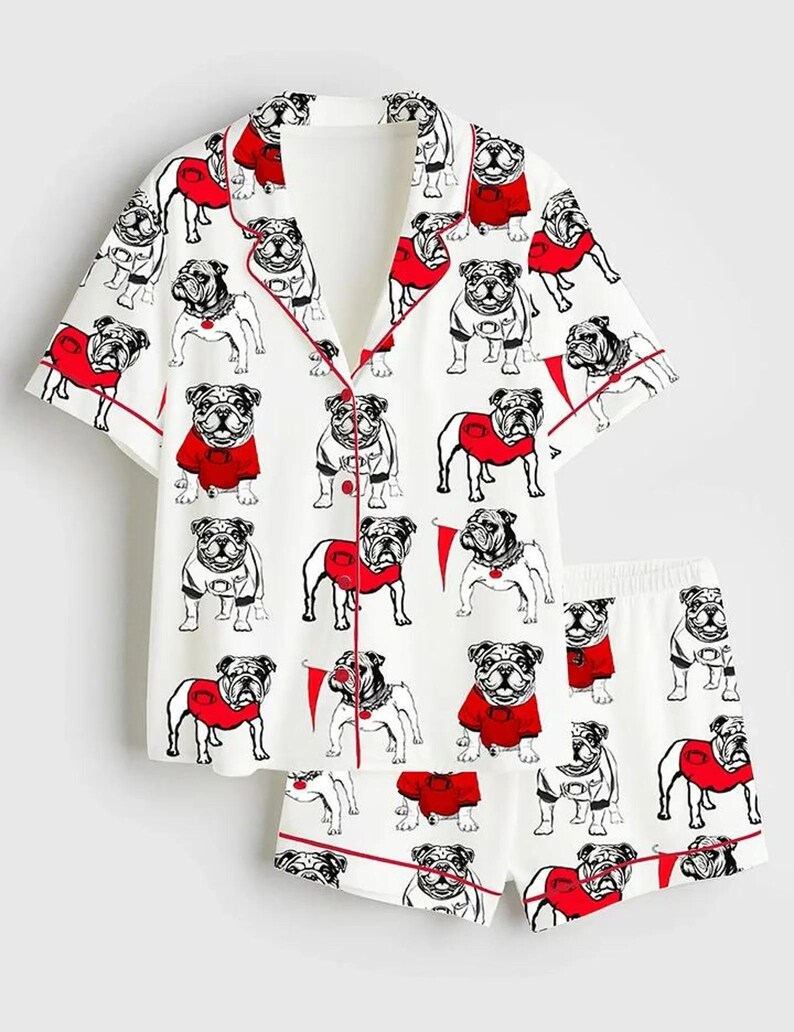 May include: A white pyjama set with a repeating pattern of bulldog illustrations in black and white, some wearing red sweaters. The set includes a short-sleeved button-up shirt and matching shorts, both trimmed with red piping.