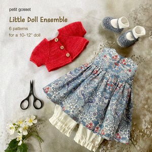 Peut inclure: Un cardigan en tricot rouge, une robe &agrave; fleurs bleu et blanc, une culotte blanche et des chaussures grises pour une poup&eacute;e de 25-30 cm. Le texte "Little Doll Ensemble" et "6 patterns for a 10-12" doll" est visible sur l'image.