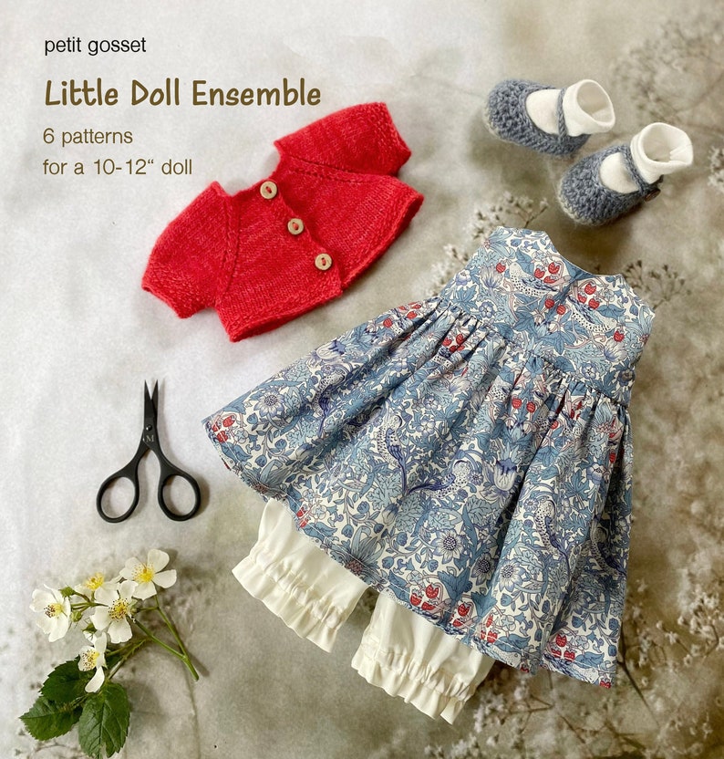 Peut inclure: Un cardigan en tricot rouge, une robe &agrave; fleurs bleu et blanc, une culotte blanche et des chaussures grises pour une poup&eacute;e de 25-30 cm. Le texte "Little Doll Ensemble" et "6 patterns for a 10-12" doll" est visible sur l'image.