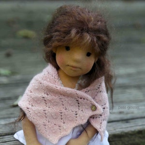 Puede incluir: Una muñeca hecha a mano con cabello castaño que lleva una chal de punto rosa con un botón. La muñeca tiene una expresión neutral y está sentada sobre una superficie de madera.