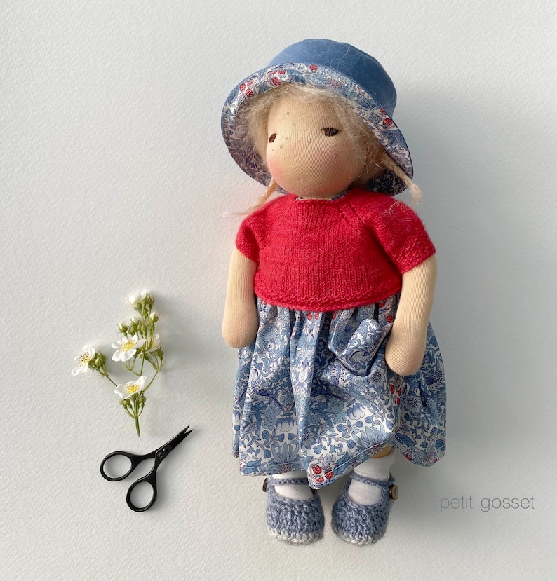 Peut inclure: Une poup&eacute;e faite &agrave; la main avec des cheveux blonds portant une robe &agrave; fleurs bleu et blanc, un pull rouge et un chapeau bleu. La poup&eacute;e porte des chaussures bleues et se tient sur un fond blanc.