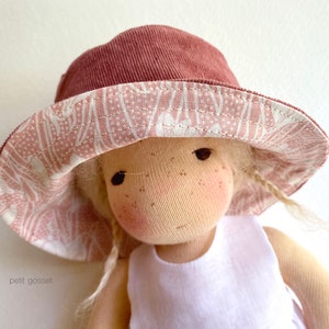 Peut inclure: Une poup&eacute;e portant un chapeau de soleil &agrave; motifs roses et blancs avec une bordure en velours c&ocirc;tel&eacute; marron. La poup&eacute;e a les cheveux blonds et porte une robe blanche sans manches.