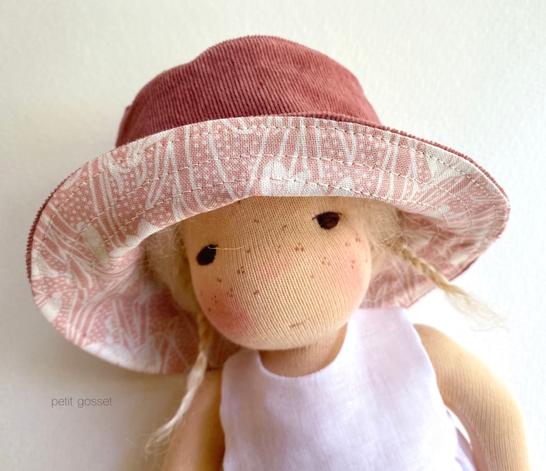Peut inclure: Une poup&eacute;e portant un chapeau de soleil &agrave; motifs roses et blancs avec une bordure en velours c&ocirc;tel&eacute; marron. La poup&eacute;e a les cheveux blonds et porte une robe blanche sans manches.