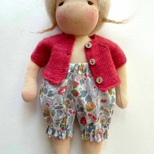 Peut inclure: Une poup&eacute;e artisanale aux cheveux blonds, portant un cardigan rouge et une culotte &agrave; fleurs. La poup&eacute;e a une expression douce et est faite de tissu doux.