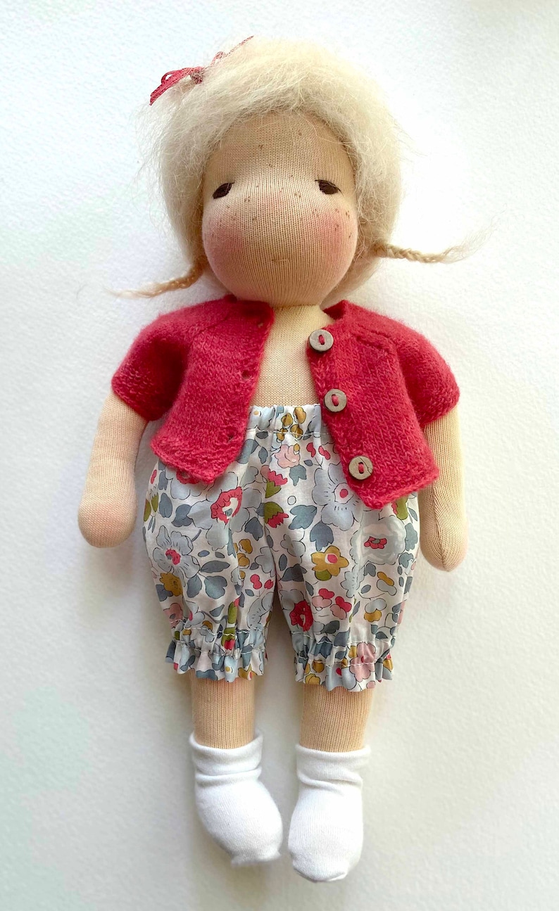 Peut inclure: Une poup&eacute;e artisanale aux cheveux blonds, portant un cardigan rouge et une culotte &agrave; fleurs. La poup&eacute;e a une expression douce et est faite de tissu doux.