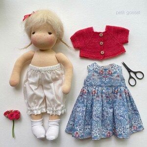 Peut inclure: Une poup&eacute;e faite &agrave; la main avec des cheveux blonds portant une culotte en coton blanc et un cardigan rouge. La poup&eacute;e tient une robe &agrave; fleurs bleu et blanc sur fond blanc.