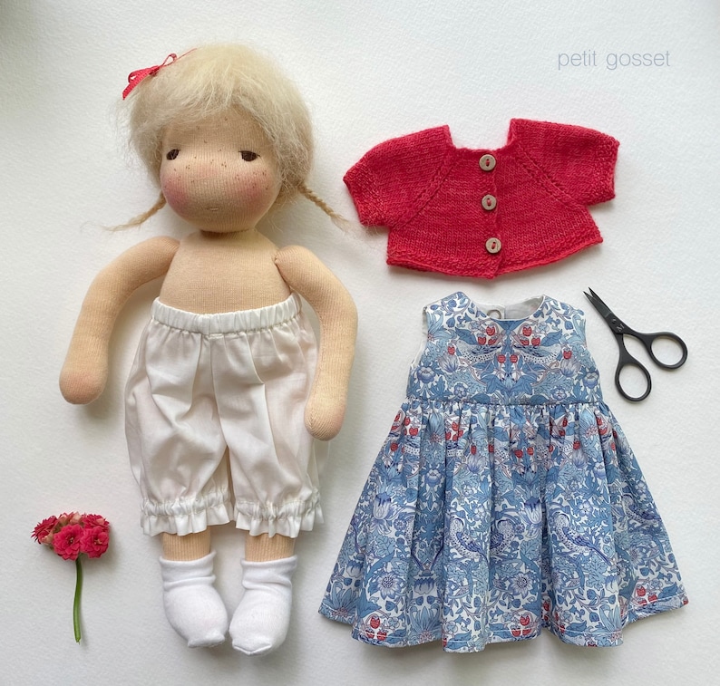 Peut inclure: Une poup&eacute;e faite &agrave; la main avec des cheveux blonds portant une culotte en coton blanc et un cardigan rouge. La poup&eacute;e tient une robe &agrave; fleurs bleu et blanc sur fond blanc.