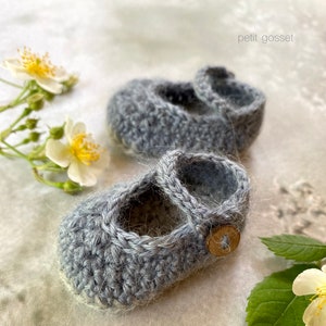 Peut inclure: Une paire de chaussons pour b&eacute;b&eacute; en crochet gris avec un d&eacute;tail bouton. Les chaussons sont sur un fond blanc avec des fleurs.