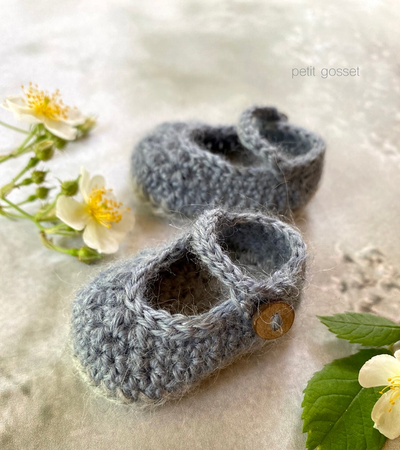 Peut inclure: Une paire de chaussons pour b&eacute;b&eacute; en crochet gris avec un d&eacute;tail bouton. Les chaussons sont sur un fond blanc avec des fleurs.