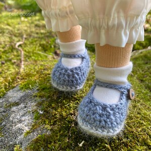 Peut inclure: Une paire de chaussures de b&eacute;b&eacute; en crochet bleu avec des bordures blanches et un bouton blanc sur une chaussure. Les chaussures sont sur un lit de mousse verte.