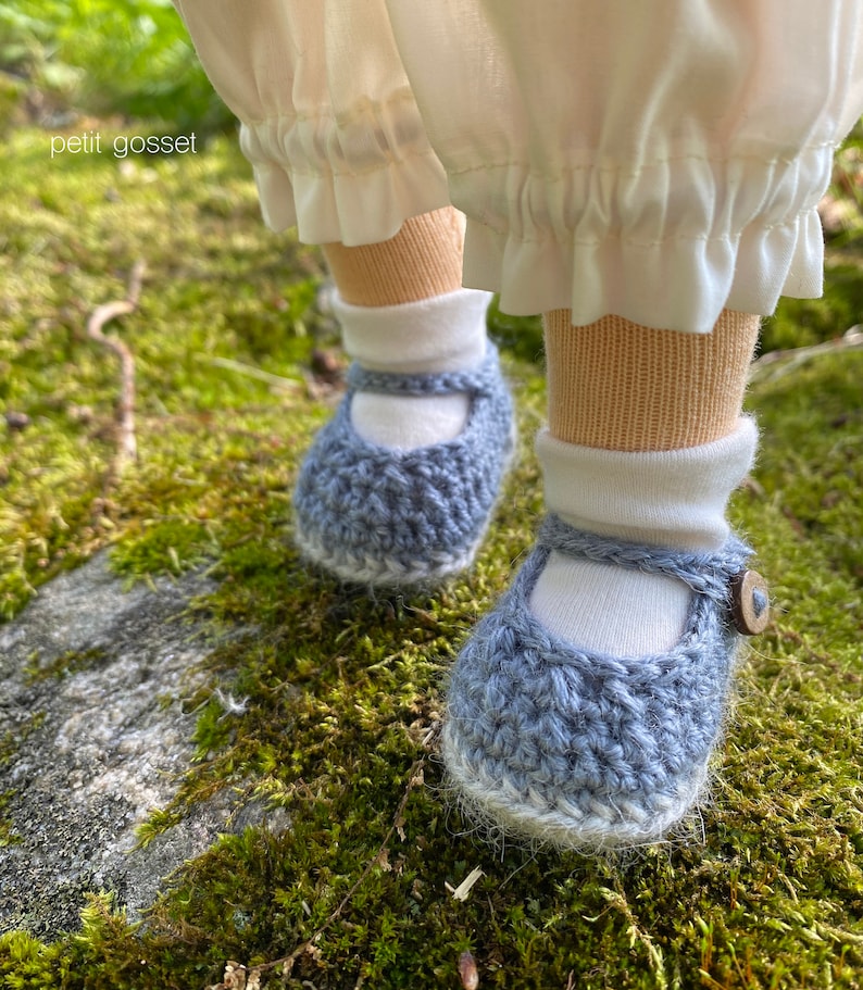 Peut inclure: Une paire de chaussures de b&eacute;b&eacute; en crochet bleu avec des bordures blanches et un bouton blanc sur une chaussure. Les chaussures sont sur un lit de mousse verte.