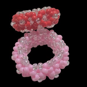 May include: Two beaded rings. The top ring is red with clear beads, and the bottom ring is pink with clear beads. The rings are made of small, round beads and are set against a black background.