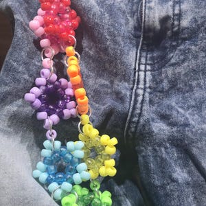 Puede incluir: Un accesorio de cuentas de colores con diseños en forma de estrella y esféricos. Las cuentas son de colores del arcoíris: rojo, rosa, morado, azul, verde y amarillo. El accesorio está sujeto a una cadena plateada.