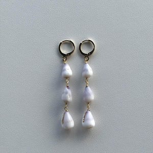 May include: A pair of gold-toned hoop earrings, each featuring a vertical drop design with three smooth, teardrop-shaped white and light purple shells suspended from the hoop by thin gold-colored metal links, creating a dangling earring set.