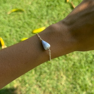 May include: A delicate gold-tone chain bracelet featuring a single, smooth, teardrop-shaped light blue stone pendant centered on the wrist. The metal chain consists of small, uniform links that connect to the polished, pale blue gemstone.