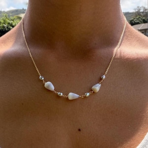 May include: A gold-toned chain necklace featuring three small, smooth, white conical shells interspersed with four tiny, round, iridescent white pearls. The delicate metal chain connects each element, creating a balanced, symmetrical arrangement around the neck.
