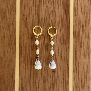 May include: A pair of gold-toned hoop earrings featuring a vertical drop design. Each earring consists of two small white oval pearls connected by gold wire, ending in a single teardrop-shaped white and grey shell charm suspended at the base.