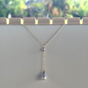 May include: A gold-toned chain necklace featuring a small, smooth, grey oval bead positioned above a larger, teardrop-shaped pendant that transitions from a dark grey top to a white base, creating a delicate lariat-style drop design.