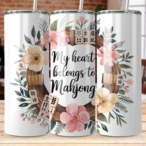 May include: White tumbler with a floral design and the text "My heart belongs to Mahjong." The design includes a wreath of flowers, leaves, and Mahjong tiles. The tumbler has a silver lid and straw.