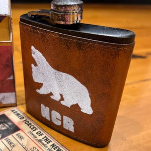 Puede incluir: Una petaca de metal forrada en cuero marrón con la silueta blanca de un oso y las letras NCR. Junto a ella hay una tarjeta de identificación rectangular titulada Armed Forces of the New California Republic y una caja Big Boss.