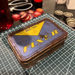 May include: A rectangular metal box with a blue lid and yellow triangle design. The box has the text "MED-TEK FIXER" in yellow. The box has a rusty, aged appearance.