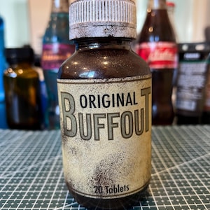 Pode incluir: Uma garrafa de vidro castanho com uma tampa branca e um rótulo que diz "ORIGINAL BUFFOUT" em uma fonte desgastada. O rótulo também indica "20 comprimidos". Outras garrafas são visíveis no fundo.