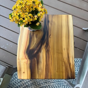 May include: A rectangular wooden side table with a natural wood grain finish. The table has a live edge on one side and is topped with a potted plant with yellow flowers. The table is approximately 24 inches long.