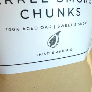 K&ouml;nnte beinhalten: Nahaufnahme eines wei&szlig;en Etiketts mit schwarzem Text, der "BOURBON BARREL SMOKE CHUNKS" lautet. Das Etikett enth&auml;lt auch "100% AGED OAK | SWEET & SMOKY" und eine Feigenillustration. Der Markenname "THISTLE AND FIG" ist unten aufgedruckt.