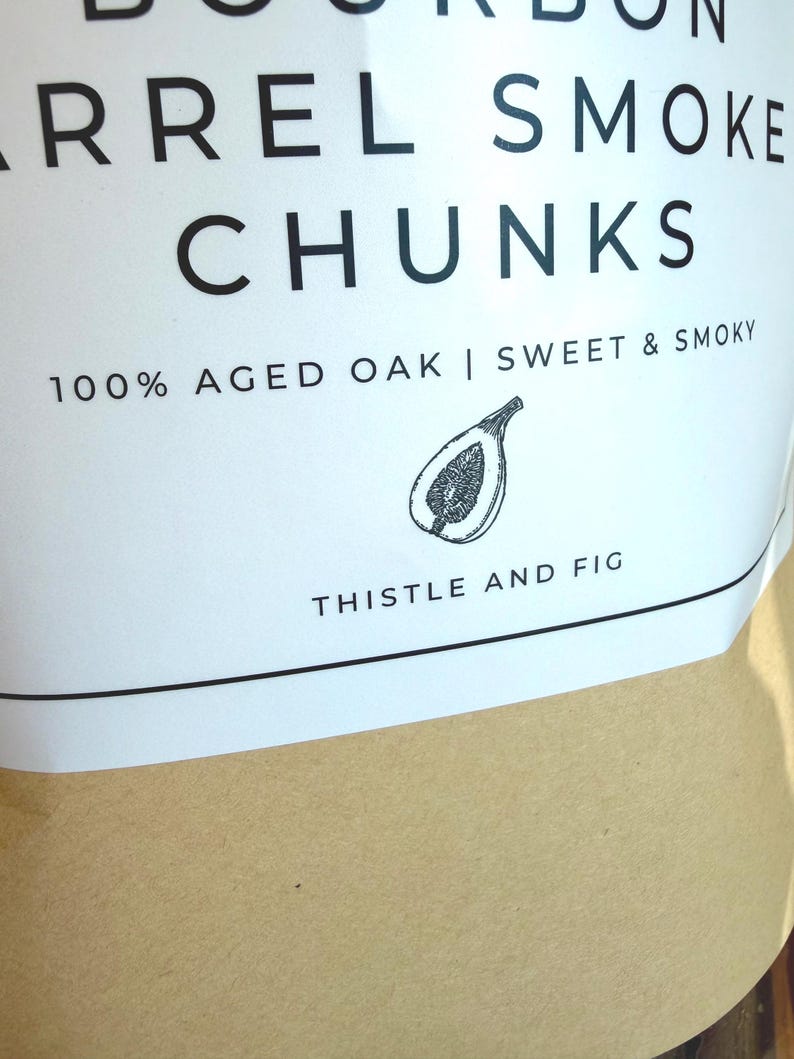 K&ouml;nnte beinhalten: Nahaufnahme eines wei&szlig;en Etiketts mit schwarzem Text, der "BOURBON BARREL SMOKE CHUNKS" lautet. Das Etikett enth&auml;lt auch "100% AGED OAK | SWEET & SMOKY" und eine Feigenillustration. Der Markenname "THISTLE AND FIG" ist unten aufgedruckt.