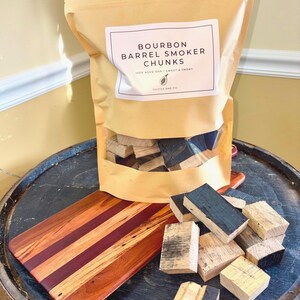 K&ouml;nnte beinhalten: Eine Packung Bourbon-Fass-R&auml;ucherst&uuml;cke mit der Aufschrift "Bourbon Barrel Smoker Chunks". Die Packung steht auf einem Holzfass mit einem Holzschneidebrett und mehreren Holzst&uuml;cken.