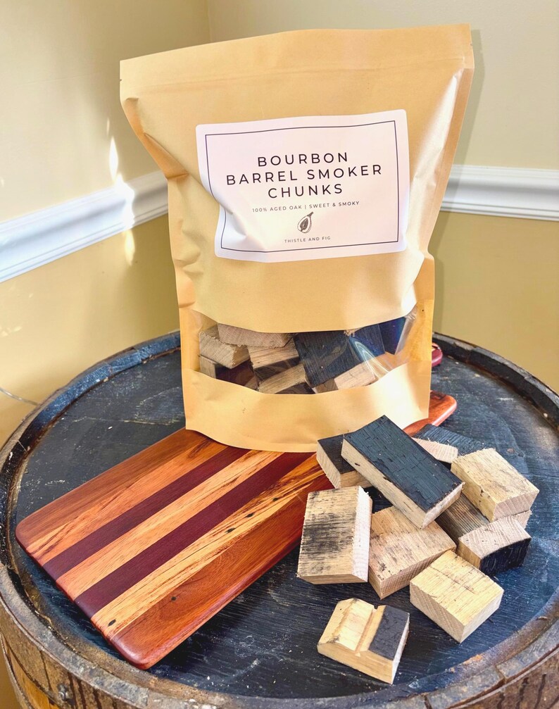 K&ouml;nnte beinhalten: Eine Packung Bourbon-Fass-R&auml;ucherst&uuml;cke mit der Aufschrift "Bourbon Barrel Smoker Chunks". Die Packung steht auf einem Holzfass mit einem Holzschneidebrett und mehreren Holzst&uuml;cken.