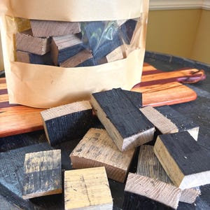 K&ouml;nnte beinhalten: Ein klarer Beutel gef&uuml;llt mit kleinen, rechteckigen Holzbl&ouml;cken, einige mit verkohlten schwarzen Oberfl&auml;chen. Die Bl&ouml;cke sind auf einer dunklen, strukturierten Oberfl&auml;che verstreut, mit einem Holzschneidebrett im Hintergrund. Das Holz scheint Eiche zu sein.