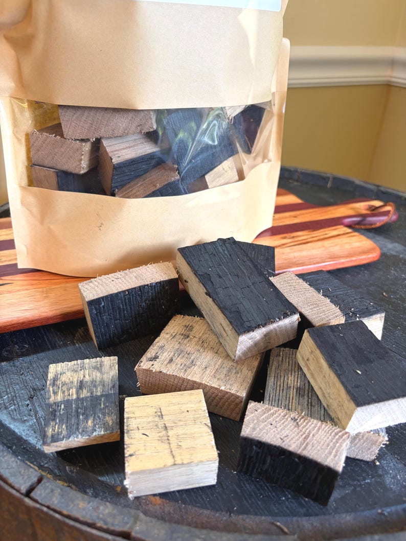K&ouml;nnte beinhalten: Ein klarer Beutel gef&uuml;llt mit kleinen, rechteckigen Holzbl&ouml;cken, einige mit verkohlten schwarzen Oberfl&auml;chen. Die Bl&ouml;cke sind auf einer dunklen, strukturierten Oberfl&auml;che verstreut, mit einem Holzschneidebrett im Hintergrund. Das Holz scheint Eiche zu sein.