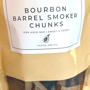 K&ouml;nnte beinhalten: Eine Packung Bourbon-Fass-R&auml;ucherchips. Das Etikett lautet "BOURBON BARREL SMOKER CHUNKS" mit dem Text "100% AGED OAK | SWEET & SMOKY" und "THISTLE AND FIG". Die Verpackung ist beige mit einem transparenten Fenster, das die Holzst&uuml;cke zeigt.