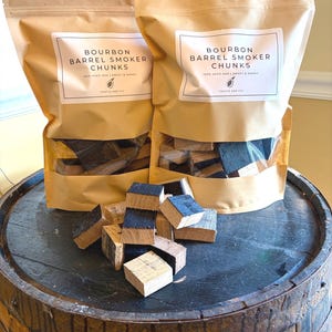 May include: Two brown paper bags of bourbon barrel smoker chunks with a clear window, sitting on a wooden barrel. The bags have white labels with black text that reads "Bourbon Barrel Smoker Chunks". Several wood chunks are scattered on the barrel.