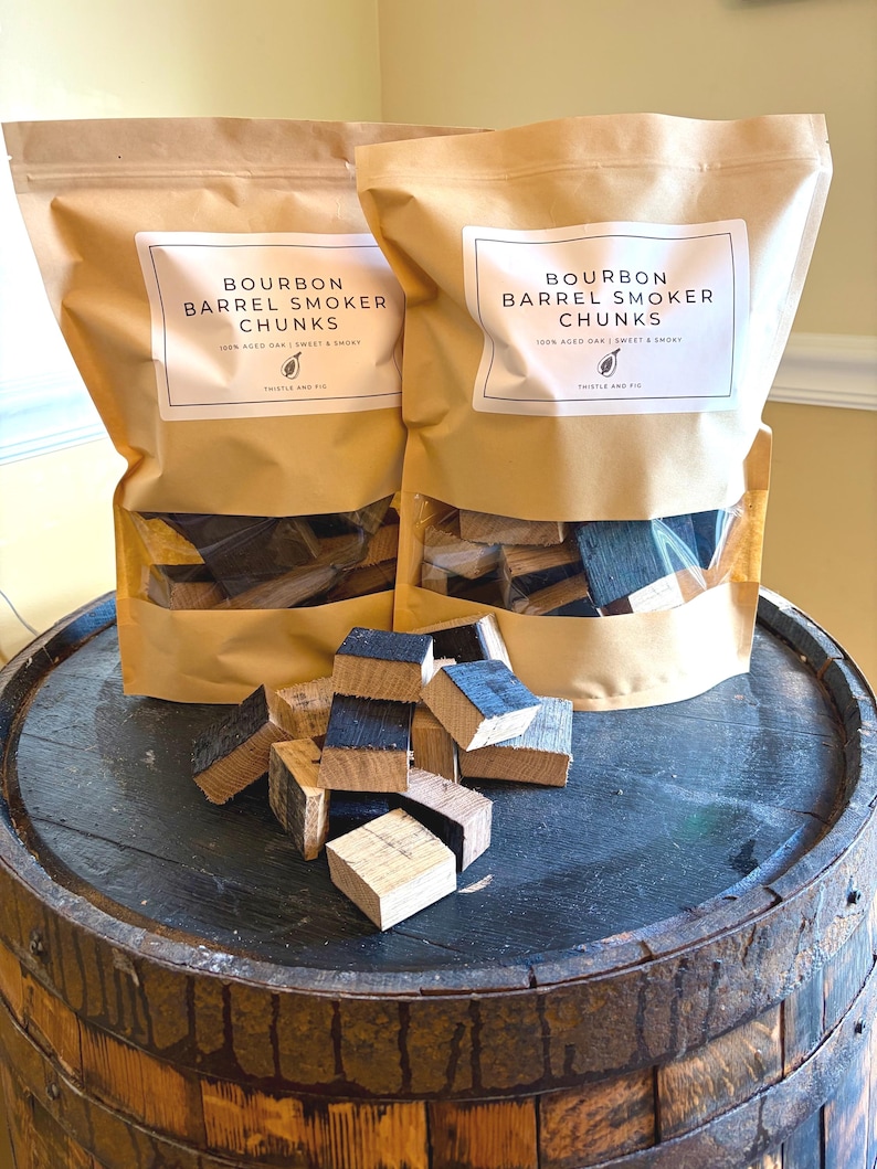 K&ouml;nnte beinhalten: Zwei braune Papiert&uuml;ten mit Bourbon-Fass-R&auml;ucherchunks mit einem transparenten Fenster, die auf einem Holzfass stehen. Die T&uuml;ten haben wei&szlig;e Etiketten mit schwarzem Text mit der Aufschrift "Bourbon Barrel Smoker Chunks". Mehrere Holzst&uuml;cke sind auf dem Fass verstreut.