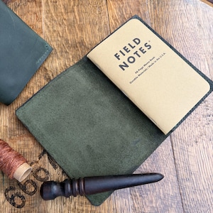 May include: A dark olive green leather notebook cover holding a tan Field Notes memo book with black text. The cover is open, revealing a textured interior. A dark brown wooden burnishing tool and a spool of copper-colored thread are nearby.