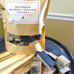 K&ouml;nnte beinhalten: Eine braune Papiert&uuml;te mit der Aufschrift "Bourbon Barrel Smoker Chunks" mit einem transparenten Fenster, das Holzst&uuml;cke zeigt. Die T&uuml;te steht auf einem dunklen Holzfass mit einem Holzschneidebrett und weiteren Holzst&uuml;cken.
