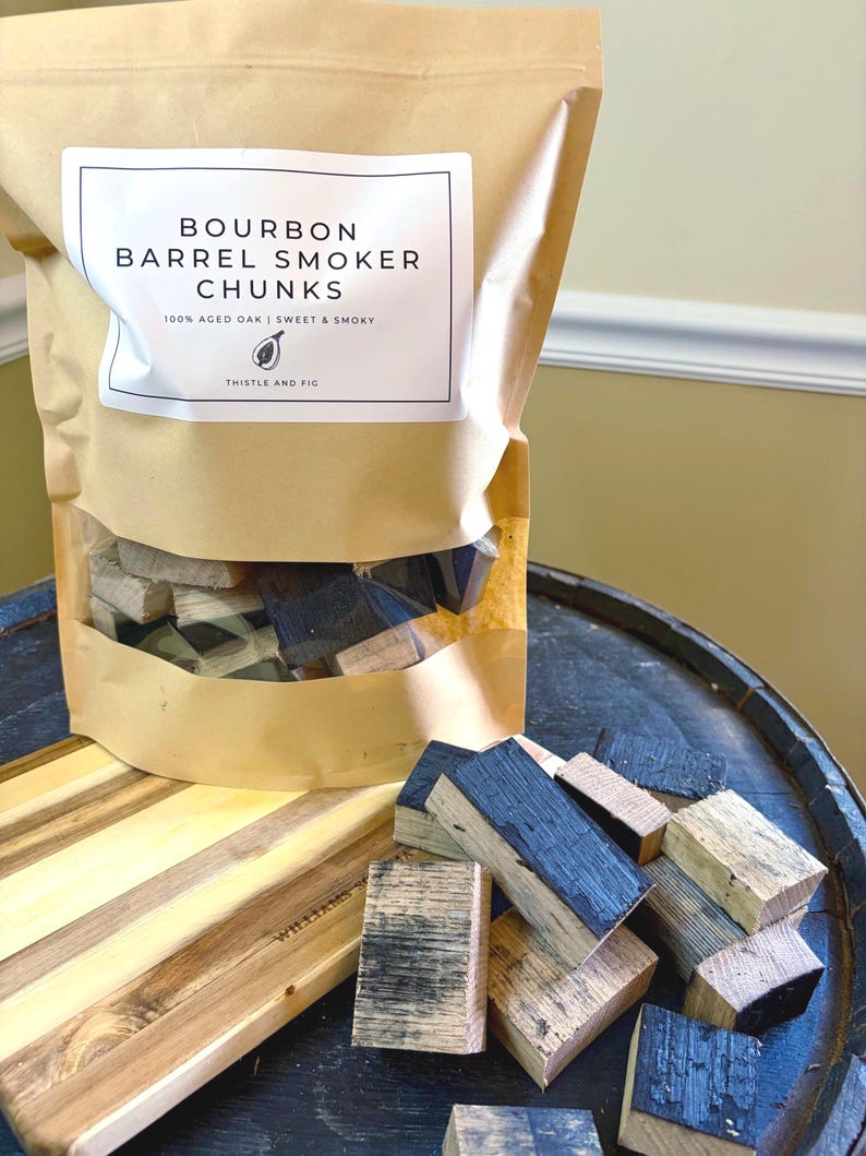 K&ouml;nnte beinhalten: Eine braune Papiert&uuml;te mit der Aufschrift "Bourbon Barrel Smoker Chunks" mit einem transparenten Fenster, das Holzst&uuml;cke zeigt. Die T&uuml;te steht auf einem dunklen Holzfass mit einem Holzschneidebrett und weiteren Holzst&uuml;cken.
