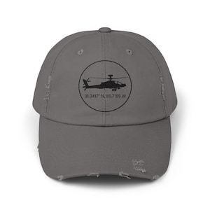May include: A distressed gray baseball cap featuring a black helicopter graphic inside a circle. The text "31.3417° N, 85.7155 W" is printed below the graphic. The cap has a curved brim and a worn, vintage look.