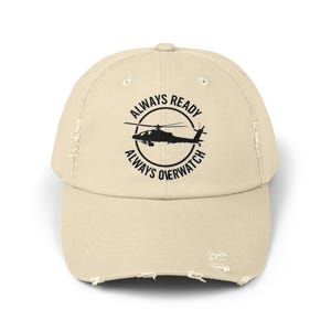 May include: A distressed beige baseball cap with a curved brim. The cap features a black graphic of a helicopter inside a circular design with the text "ALWAYS READY" and "ALWAYS OVERWATCH".